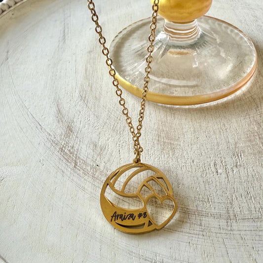 May include: A gold volleyball necklace with a heart cut-out. The pendant is engraved with the name &#39;Ariza&#39; and the number &#39;#8&#39;.
